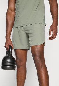 Žalios sportinės šortai, pagaminti iš lengvos medžiagos su lygia tekstūra. Juodas kettlebell'as laikomas dešinėje rankoje, demonstruojantis matinį paviršių.