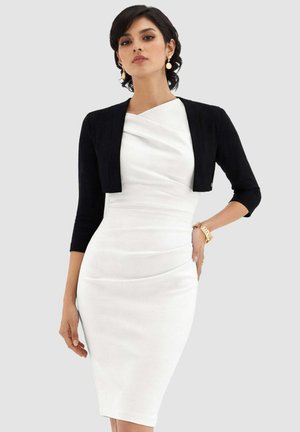 Donna che indossa un abito bianco aderente con una giacca corta nera, orecchini d'oro e un braccialetto d'oro in piedi davanti a uno sfondo semplice.