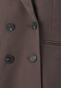Manteau croisé marron foncé avec quatre boutons noirs et une poche passepoilée horizontale sur le côté droit.