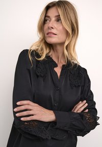 Blusa negra con detalles de encaje en los hombros y mangas, con botones en el frente, cuello con volantes y mangas largas. Tejido liso y ligeramente brillante.