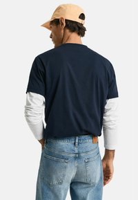 Tumesinine lühikeste varrukatega t-särk, millele on selga pandud valge pika varrukaga särk, koos helesiniste denimitega, mille taga on pruun nahast silt.