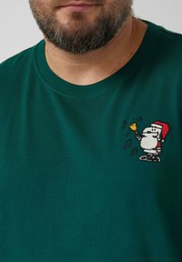 Grünes Baumwoll-T-Shirt mit rundem Ausschnitt, bestickt mit einem Weihnachtsmann, der eine Glocke hält, umgeben von musikalischen Noten.