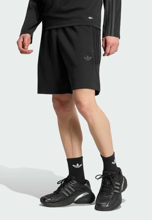 Męskie nogi ubrane w czarne spodenki Adidas, czarne skarpetki Adidas i czarne buty sportowe, stojące na jasnoszarym tle.