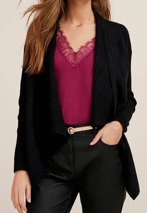 Femme portant un cardigan noir côtelé sur un haut violet orné de dentelle, avec un pantalon noir et une ceinture beige clair, main dans la poche.