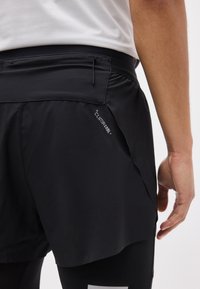 Gros plan d'une personne portant un short Climacool+ noir par-dessus des leggings noirs et une chemise blanche, montrant une poche arrière zippée.