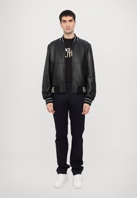 Jachetă bomber din piele neagră cu manșete ribbed cu dungi albe și tiv în contrast cu logo, purtată peste un tricou grafic negru și pantaloni închiși la culoare.