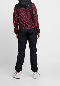 Czerwona i czarna kurtka wiatrówka z wzorem, z czarnym kapturem i elastycznymi mankietami, w połączeniu z czarnymi lekkimi spodniami. Całość dopełniają białe sneakersy.