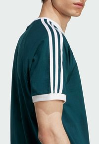 T-shirt vert foncé avec des accents rayés blancs sur les épaules et des poignets côtelés blancs, avec un design à col rond classique. Texture de tissu lisse.