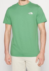 Green cotton t-shirt with a crew neck and short sleeves. Features a small white logo on the left chest. Smooth texture, casual design.
