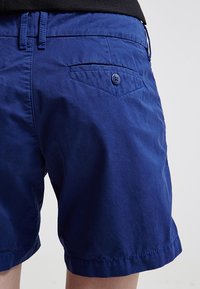 Detailní záběr na osobu v modrých šortkách s knoflíkovou zadní kapsou a poutky na opasek, na světlém jednobarevném pozadí.