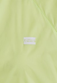 Svetlo zelena tkanina z drobno teksturo, ki se ponaša z belim logotipom Levi's na sredini, obdanem z gladkim, rahlo sijočim materialom.