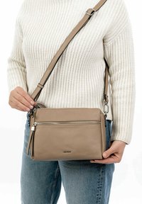 Beige leather crossbody bag with a front zip pocket, adjustable strap, and smooth texture. The bag is worn on a person’s shoulder.