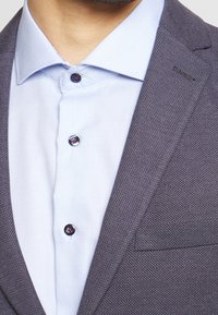 Szary blazer o fakturowanej powierzchni z jasnoniebieską koszulą z kołnierzykiem pod spodem, posiadający czarne guziki z czerwonymi akcentami na koszuli.