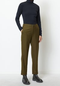Grüne Wollhose mit geradem Schnitt, mit Knopfverschluss und seitlichen Taschen, kombiniert mit einem eng anliegenden schwarzen Rollkragenpullover und schwarzen Stiefeletten.