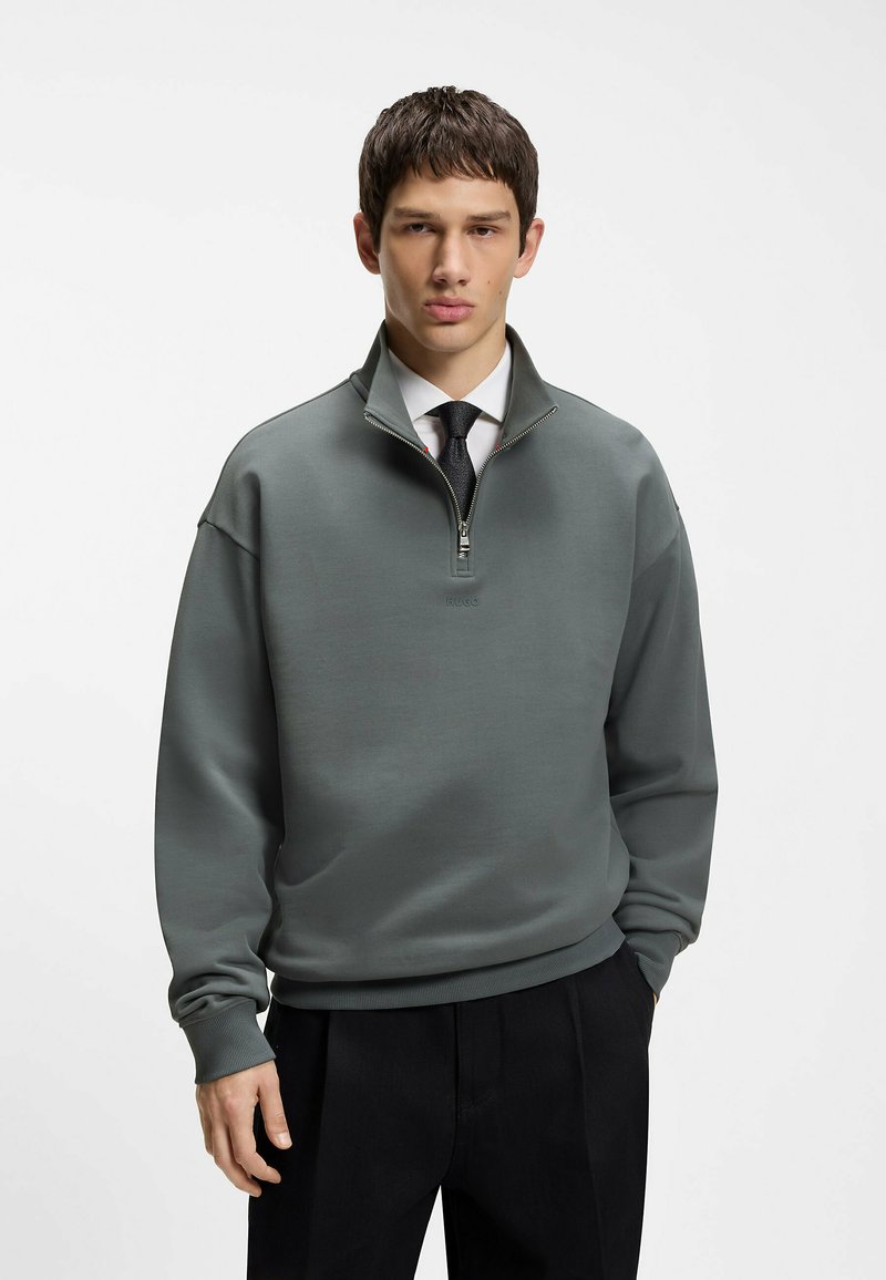 Jeune homme portant un sweat-shirt gris à fermeture éclair sur un t-shirt blanc et une cravate noire, avec un pantalon noir, debout devant un fond blanc uni.