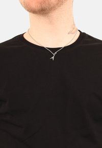 Collar plateado con un colgante geométrico, llevado sobre una camiseta negra de cuello redondo, que presenta una cadena delgada y un diseño intrincado.