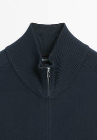 Granatowy sweter dzianinowy z prążkowanym, wysokim kołnierzem i częściowo zasuniętym srebrnym zamkiem błyskawicznym na środku przedniej części.