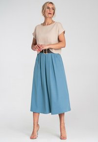 Beżowa bluzka z krótkim rękawem o luźnym kroju, zestawiona z niebieskimi spódnicami w plisy do połowy łydki, podkreślona czarnym paskiem i cielistymi sandałami na obcasie.