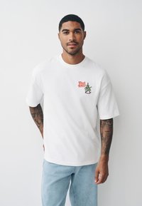 Balta kokvilnas T-krekls ar īsām piedurknēm, uz kura ir krāsains grafiskais elements "Deck the Halls" un karikatūrisks Ziemassvētku eglītes attēls kreisajā krūtīs.