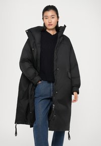 Long manteau matelassé noir avec un col haut et une fermeture éclair, doté de poches latérales et d'une texture lisse et brillante. Porté sur un pull noir.