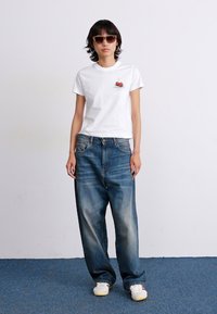 Camiseta de algodón blanca con gráfico de cereza, corte holgado, combinada con jeans de mezclilla azul sueltos. El modelo lleva gafas de sol y calzado blanco.