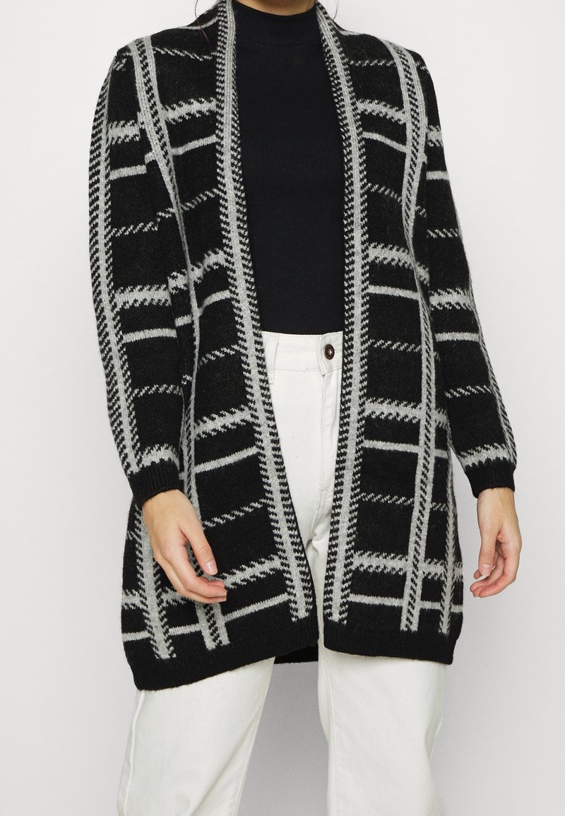 Femme portant un cardigan à motifs noir et gris ouvert sur un haut noir et un pantalon blanc, debout devant un fond uni.