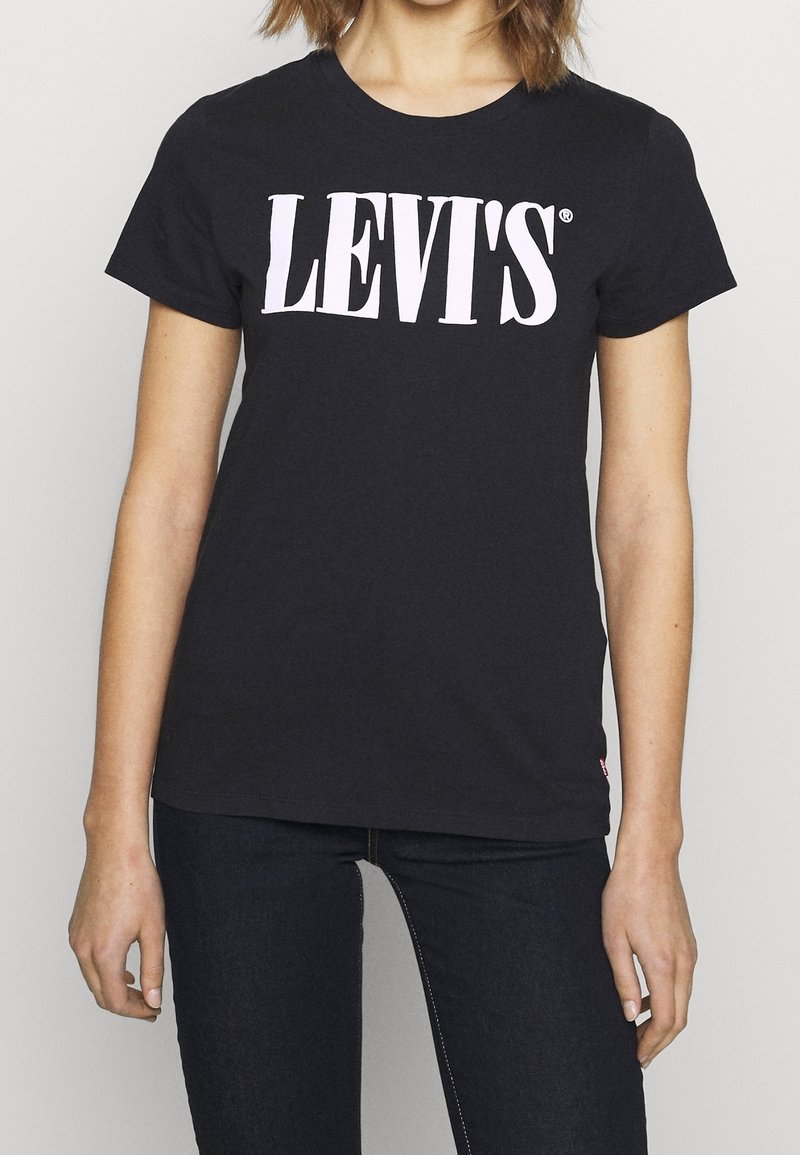 Femme portant un t-shirt noir Levi's avec un grand logo blanc et un jean foncé, debout devant un fond uni.