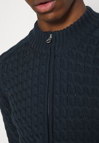 Närbild av en person som bär en mörkblå kabelstickad zip-up-tröja med en ribbad krage och synlig dragkedja.