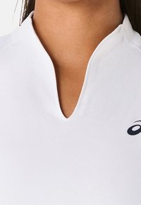Top atlético blanco con cuello alto, que presenta un escote en forma de V corto. La tela es suave, ligera y cuenta con un pequeño logo en azul marino.