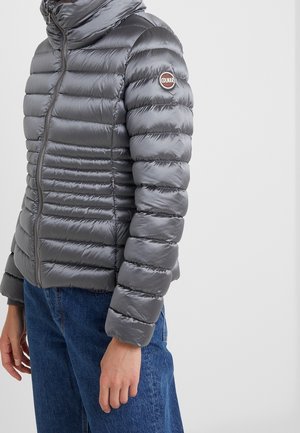 Persona vistiendo una chaqueta acolchada brillante gris con costuras horizontales y un parche con el logo de Colmar en la parte superior del brazo, combinada con jeans azules.