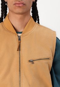 Giacca bomber color sabbia con zip frontale, colletto a costine e tasca con zip sul petto a sinistra. Il tessuto appare testurizzato con una leggera lucentezza.