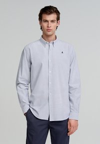 Button-up-Hemd mit blau-weißem Streifenmuster, Kragen, langen Ärmeln und einem kleinen Logo auf der linken Brust. Der Stoff wirkt glatt.