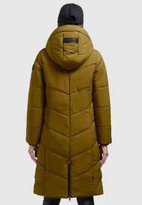 Persona con abrigo largo de plumas verde oliva con capucha, vista desde atrás, también lleva una gorra negra y pantalones negros.