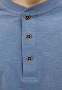 Világoskék henley ing, textúrált anyagból készült, három sötétbarna gombbal és lekerekített nyakrésszel. Egyszerű varrási részletezés.