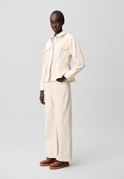 Chaqueta vaquera color crema con dos bolsillos frontales y cintura ajustada, combinada con pantalones de pierna ancha y zapatos marrones con cordones.