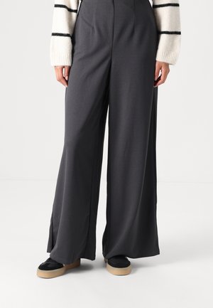 Pantaloni a gamba larga in tessuto grigio scuro, con una texture liscia e spacchi laterali. Abbinati a sneakers nere e a un maglione a righe bianche.