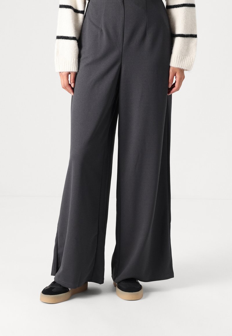 Pantalons larges en tissu gris foncé, avec une texture lisse et des fentes latérales. Associés à des baskets noires et un pull rayé blanc.