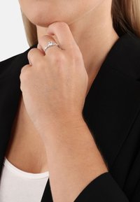 Anillo de plata con una piedra central clara, acentuado por piedras más pequeñas en una banda texturizada. Mano posicionada cerca del mentón, vistiendo un blazer negro.