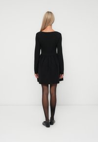 Vestido negro con mangas largas, cuerpo ajustado y falda acampanada. Tejido suave, escote redondeado y se lleva con zapatos planos negros y medias transparentes.