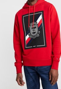 Hombre wearing un hoodie rojo con un emblema de escudo negro, blanco, rojo y azul y las letras "TH" en el pecho, combinado con jeans azul oscuro.
