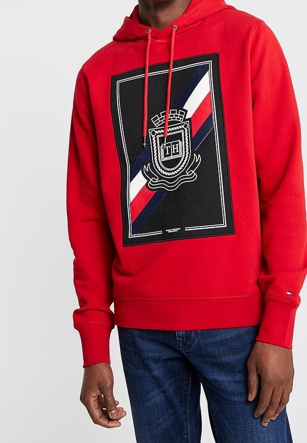 Hombre wearing un hoodie rojo con un emblema de escudo negro, blanco, rojo y azul y las letras "TH" en el pecho, combinado con jeans azul oscuro.