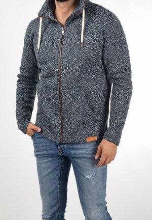 Man wearing a dark gray zip-up hoodie with white drawstrings and blue ripped jeans, standing with one hand in pocket against a white background.