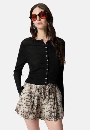 Donna con capelli lunghi e scuri indossa occhiali da sole oversize con montatura tartarugata, un cardigan nero con bottoni e pantaloncini beige con fantasia effetto pelle di serpente e coulisse.