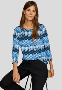 Blau gestreiftes Langarmoberteil mit Rundhalsausschnitt. Es hat Streifen in verschiedenen Blautönen und Weiß. Kombiniert mit schwarzen Hosen.