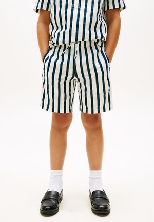 Bambino che indossa pantaloncini e camicia a righe verticali blu e bianche, calzini bianchi e scarpe slip-on nere, in piedi con le mani in tasca.