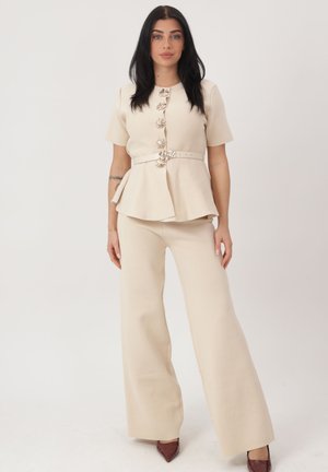 Donna con capelli scuri che indossa una maglia peplum beige a maniche corte con cintura, bottoni decorativi e pantaloni a gamba larga abbinati, in piedi su sfondo bianco.