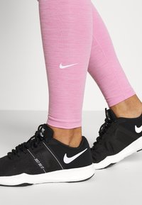 Růžové sportovní legíny s texturovaným povrchem a bílým logem Nike, spárované s černými teniskami s mesh designem a bílými akcenty.