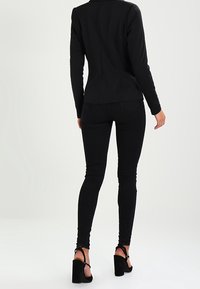 Svart figurnära kavaj med långa ärmar, i kombination med svarta skinny jeans och högklackade skor, sedd bakifrån. Slät tygstruktur.