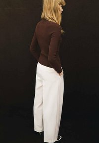 Maglione marrone a costine abbinato a pantaloni bianchi a palazzo, visto da dietro. Capelli lunghi e mani nelle tasche. Sfondo nero.