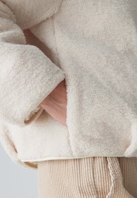 Kremowa, puszysta kurtka o miękkim, teksturowanym wykończeniu z polaru, z widoczną ręką w kieszeni i gładkim wykończeniem mankietów.